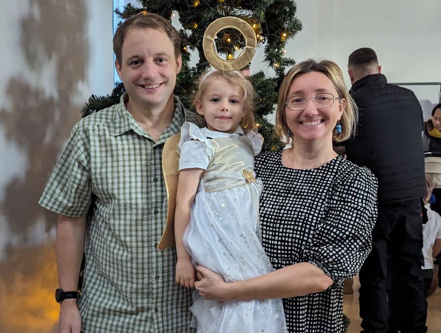 A family photo with Geoffrey Hayward wearing a green plaid shirt, his youngest child Lizzie in a white angel costume with golden wings and a halo, and Ewa Hayward wearing a black and white patterned dress. They are standing in front of a Christmas tree, smiling at the camera, with other people and holiday decorations blurred in the background. The photo was taken after Lizzie's school nativity show.