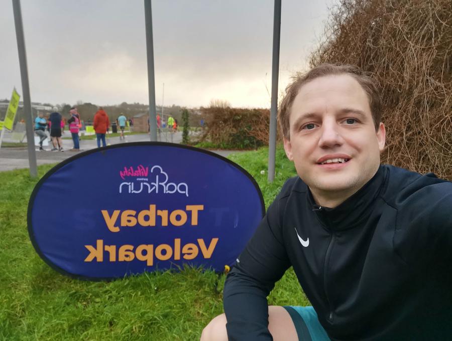 Geoffrey Hayward seated next to the Velopark Torbay parkrun sign, capturing a selfie. He is clad in a black sports outfit and appears content. The backdrop features a cloudy sky with other participants and volunteers visible in a park environment.