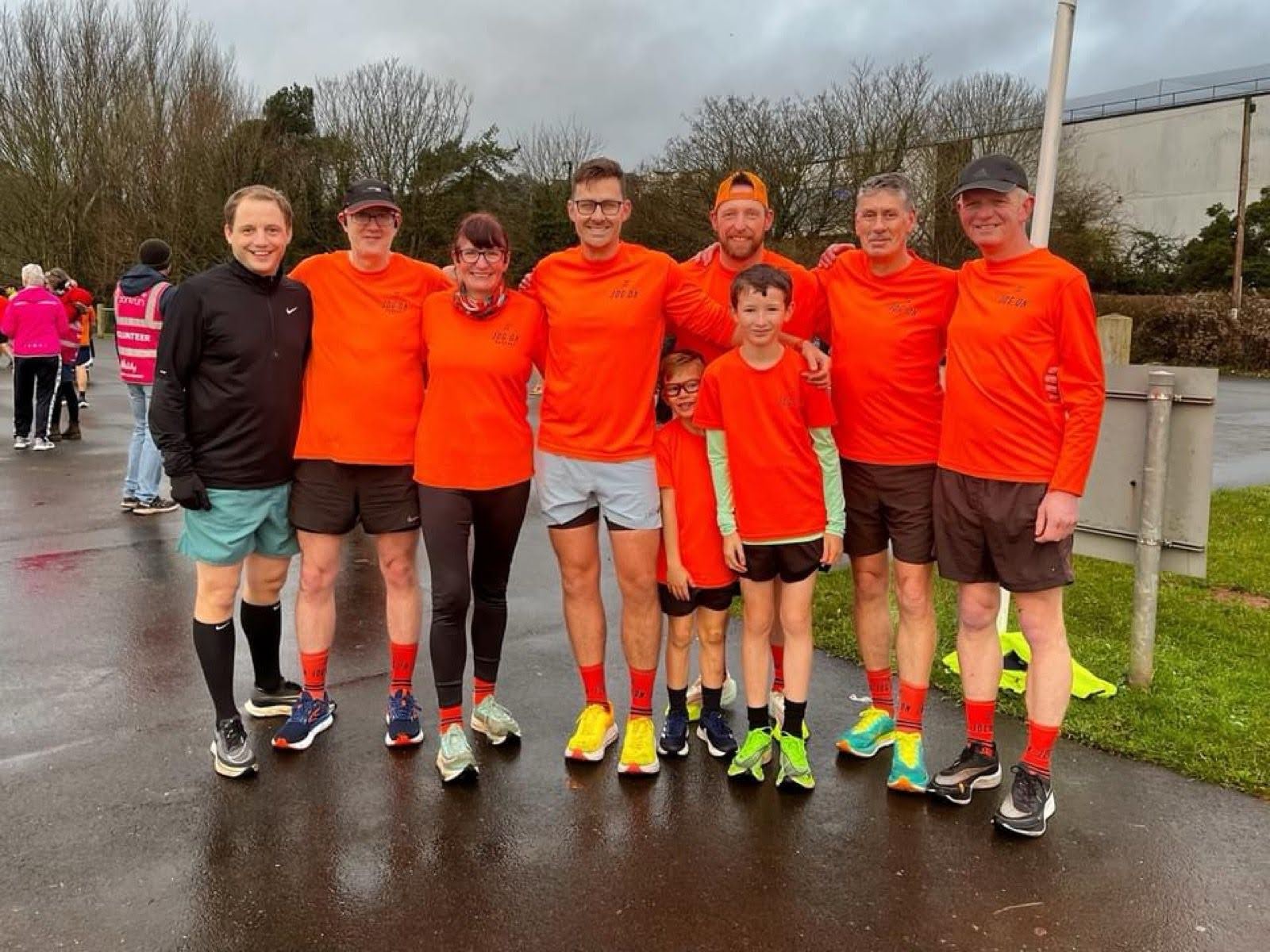 A group photo of seven adults and two children, with most wearing bright orange ‘Jog On’ tops. They are standing on a wet pavement at the Velopark Torbay as part of a running crew. The individual not in orange is wearing a black top and is positioned on the left.
