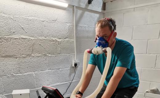 Geoffrey Hayward undergoing a cardio workout on a stationary bike inside a room with white walls, connected to a VO2 Max testing apparatus with a breathing mask, surrounded by exercise equipment.